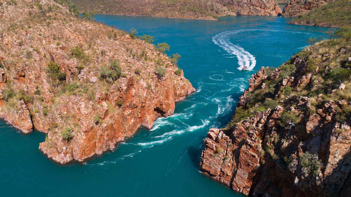 Turquoise waters swirl between rugged cliffs, with a boat leaving a wake as it navigates the scenic landscape. Lush greenery contrasts with the reddish rock formations.