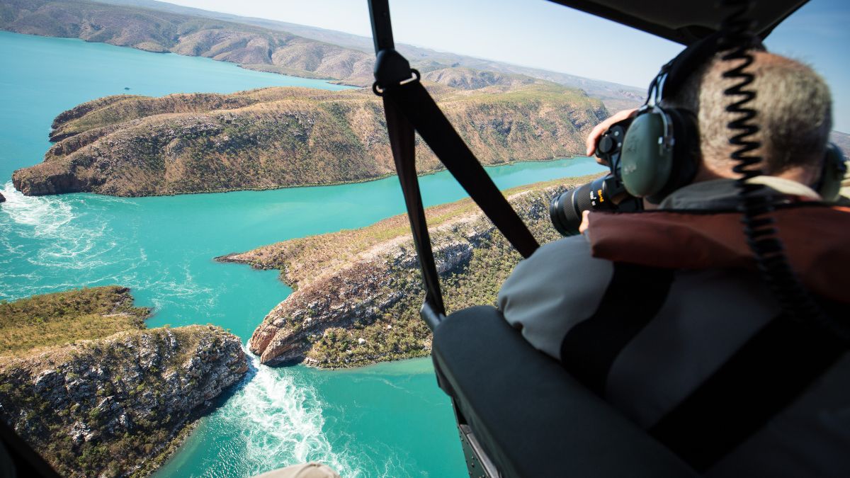 A person in a helicopter takes photographs of a stunning coastal landscape with turquoise waters, rocky cliffs, and winding shorelines.