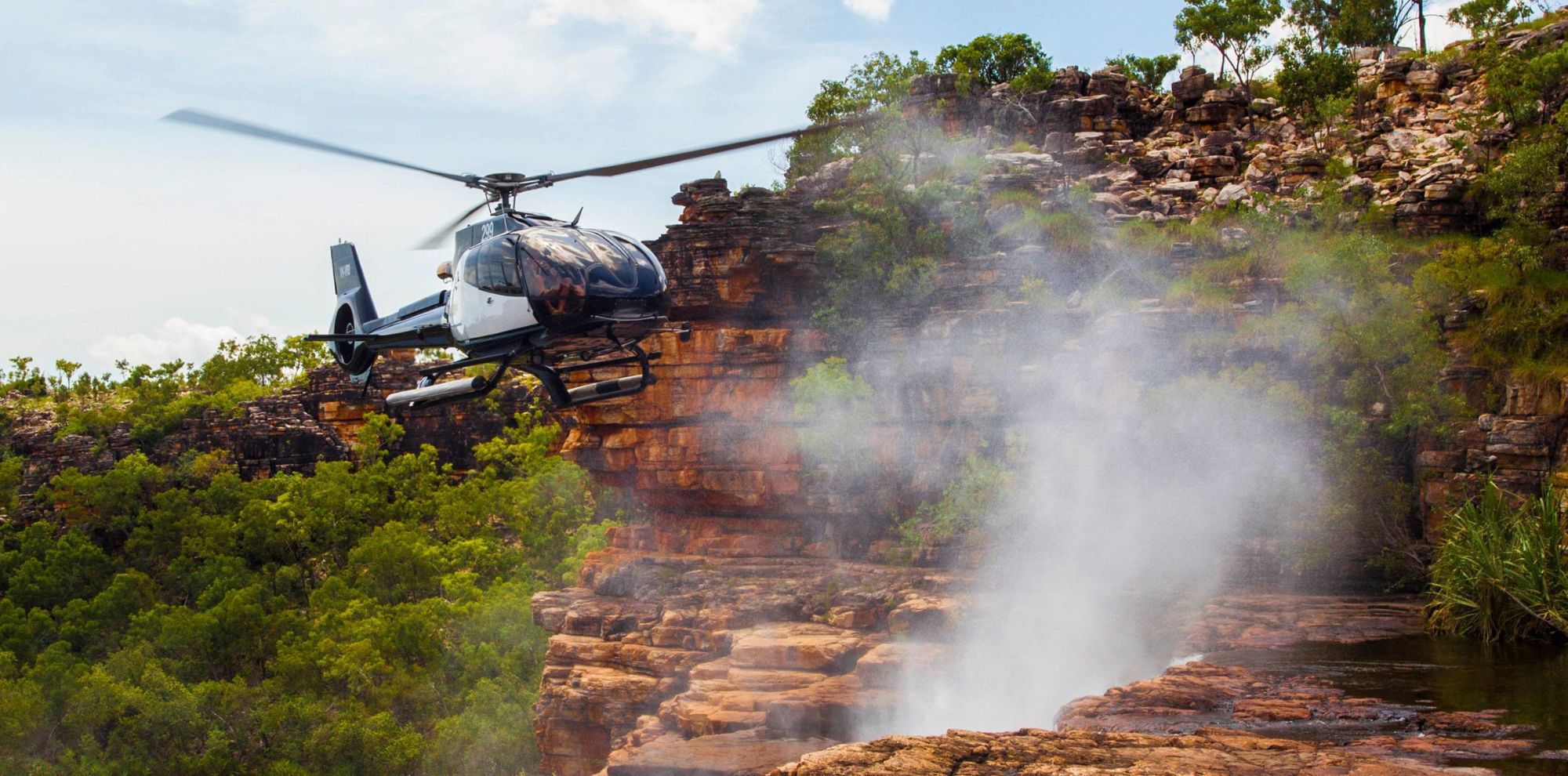A helicopter hovers near a rocky cliff with water cascading down, creating a misty spray. Lush greenery surrounds the area, adding to the natural landscape.