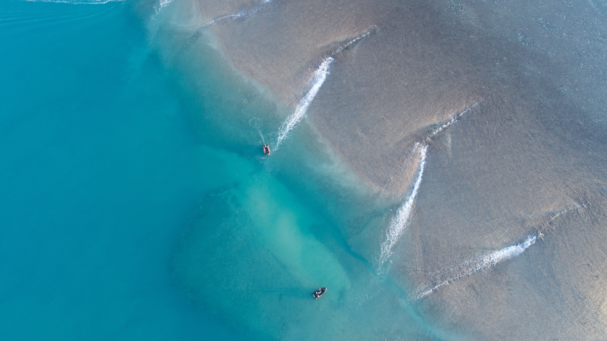 Two jet skiers glide across a bright turquoise sea, creating waves and ripples in the clear water. The sandy seabed is visible beneath the surface, adding to the vibrant scene.