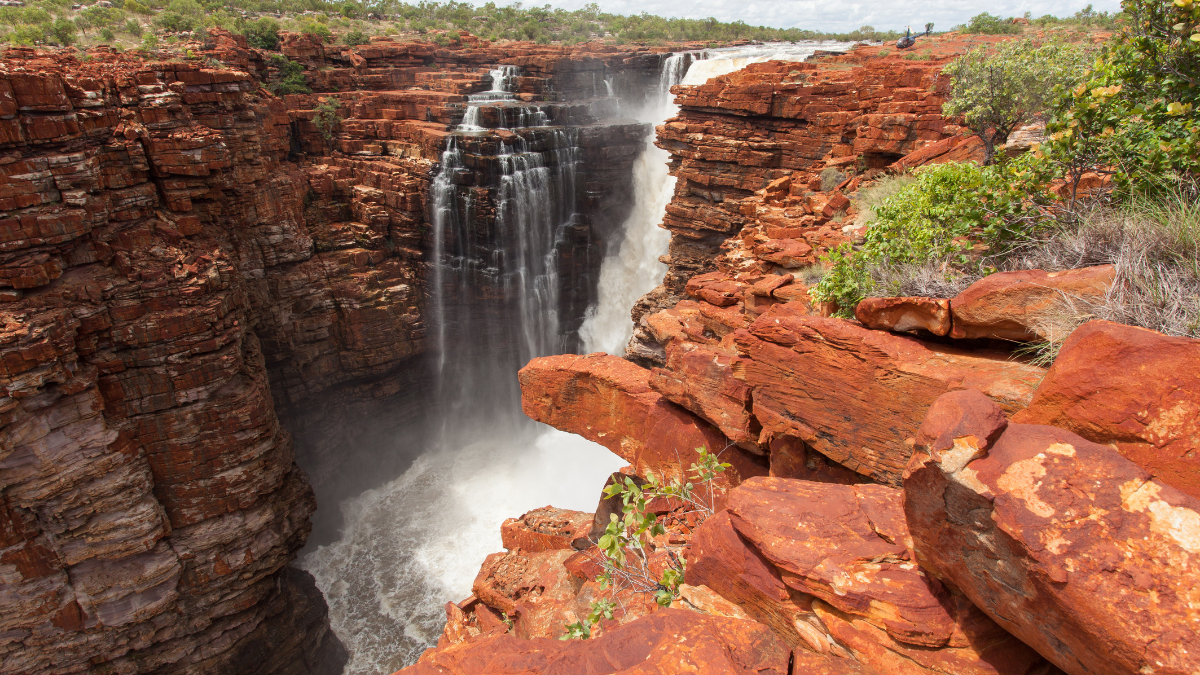 A stunning waterfall cascades over rugged red rock cliffs, surrounded by lush greenery. The powerful water plunges into a deep gorge, creating a misty spray.