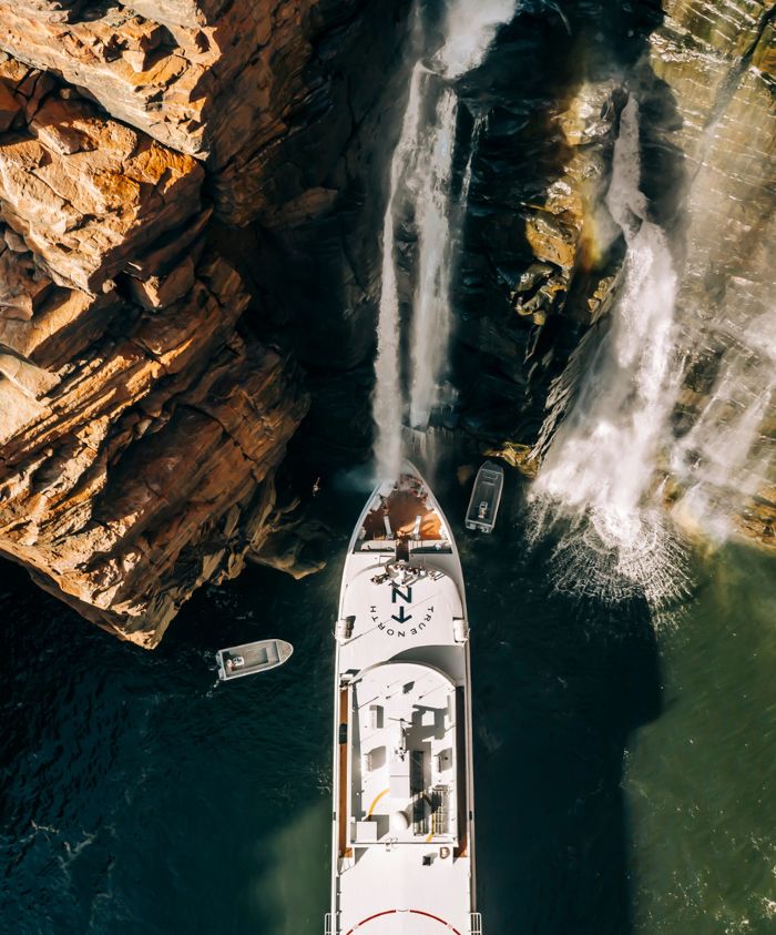 Aerial view of a large boat positioned near a rocky cliff, with waterfalls cascading down into the water. Smaller boats are visible nearby.