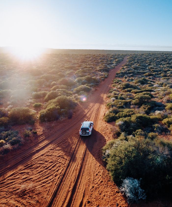 Aerial view of a car driving along a red dirt track through a vast landscape of scrub and low vegetation, with the sun rising in the background.
