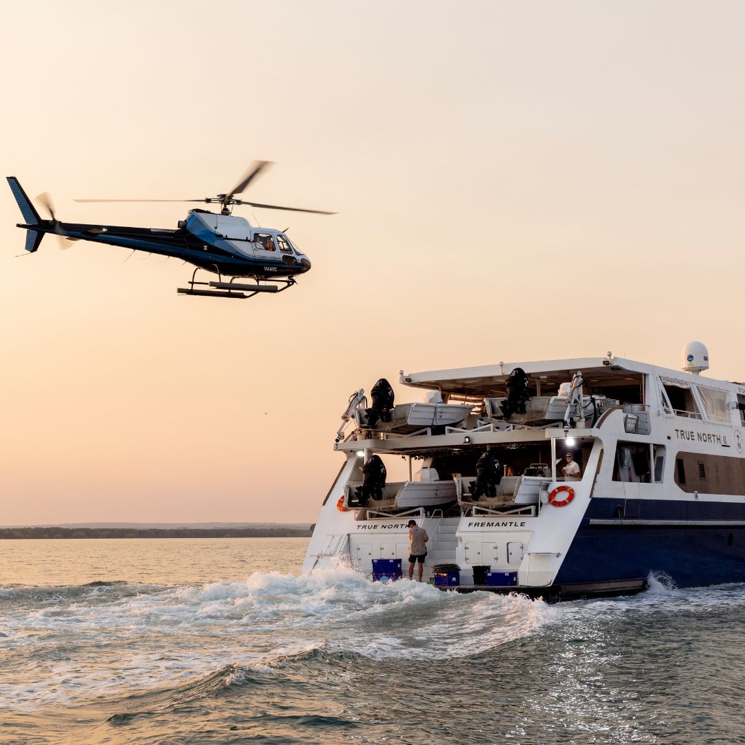 A helicopter hovers above a large boat named "True North", which is moving through calm waters at sunset. The scene captures a dynamic interaction between the helicopter and the boat.