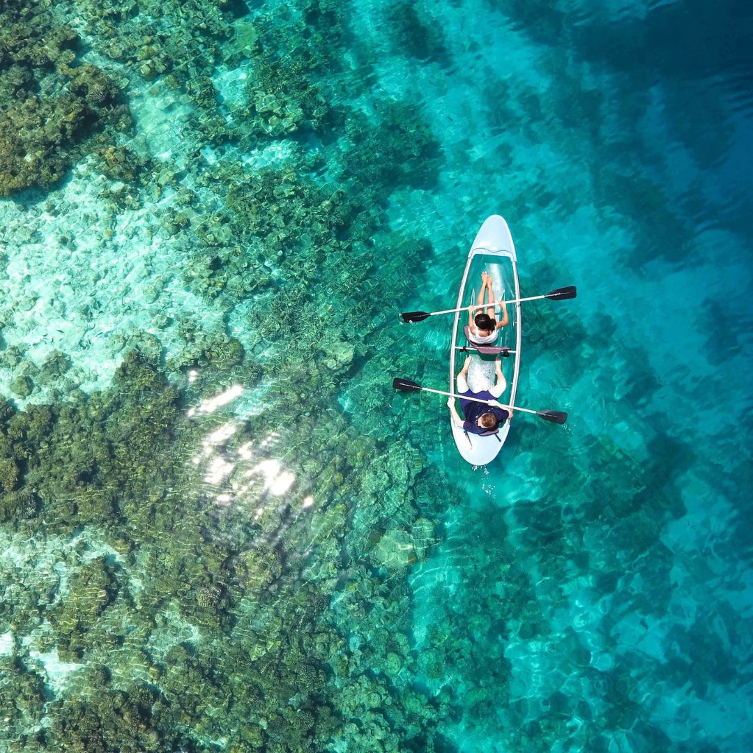 Two people paddle a kayak over clear, turquoise water, revealing vibrant coral reef below. Sunlight sparkles on the surface, enhancing the serene atmosphere.