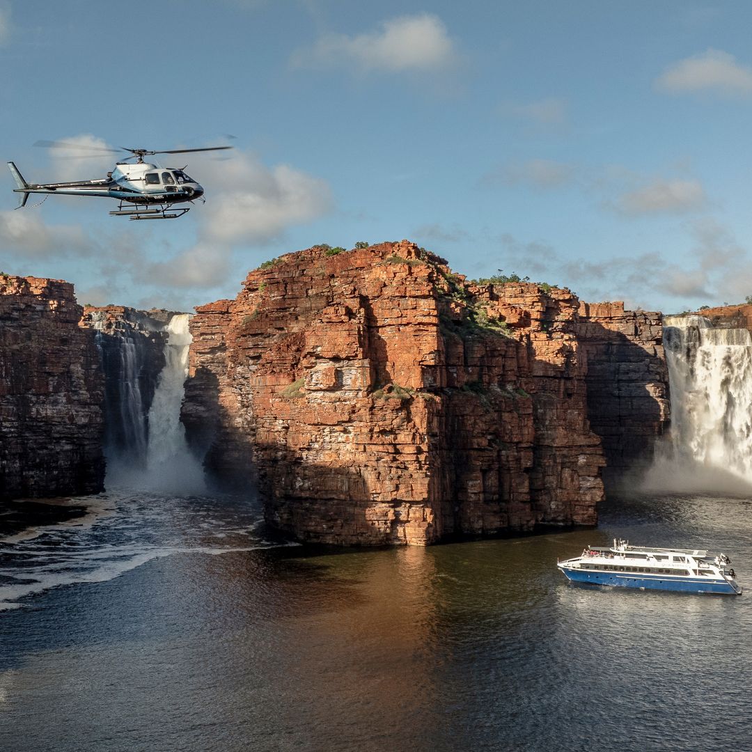 A helicopter flies over a rocky landscape with waterfalls cascading down into a river. A boat is anchored nearby, surrounded by towering cliffs and lush greenery.