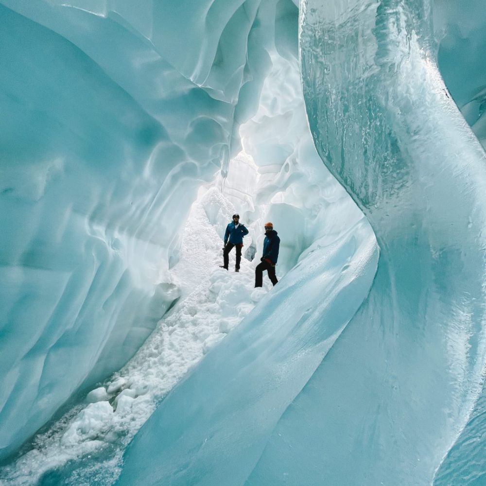 Two people stand inside a majestic ice cave, surrounded by bright blue ice formations and glistening snow. The light filters through the ice, creating a stunning, ethereal atmosphere.