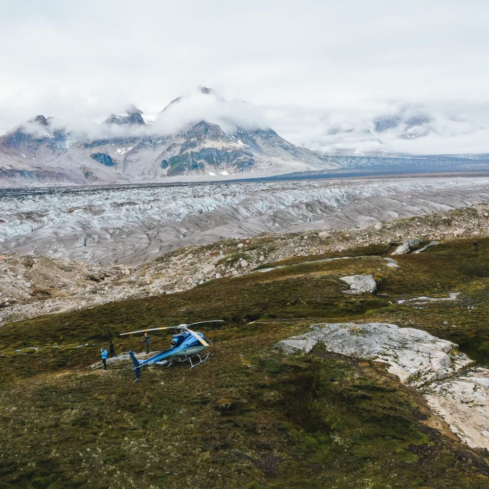 A blue helicopter sits on green terrain near a glacier, with majestic mountains and cloudy skies in the background.