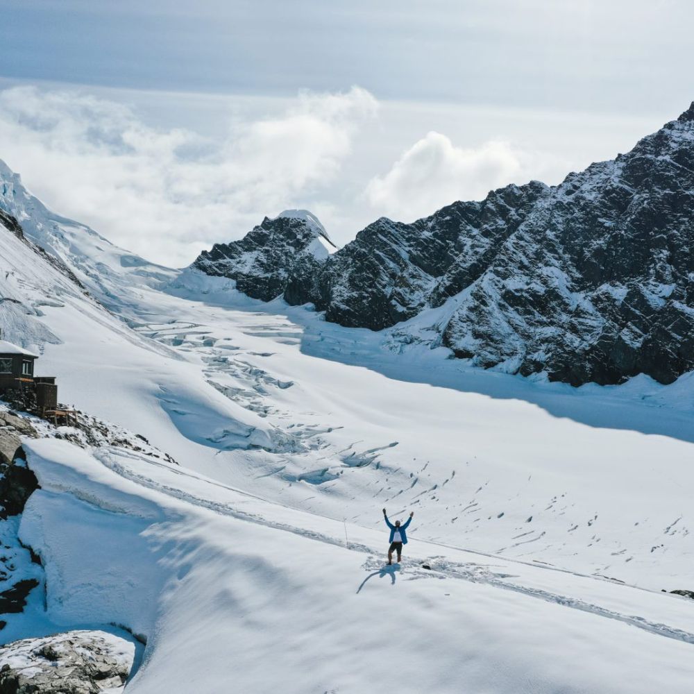 A person stands with arms raised on a snow-covered glacier, surrounded by towering mountains and a bright sky.