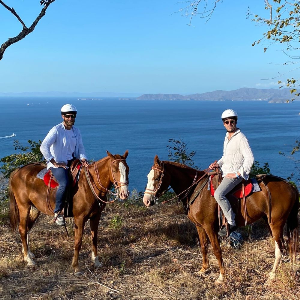 Two riders on brown horses, wearing helmets, pose on a hillside overlooking the blue ocean and distant mountains. The sun shines brightly, and trees frame the scene.