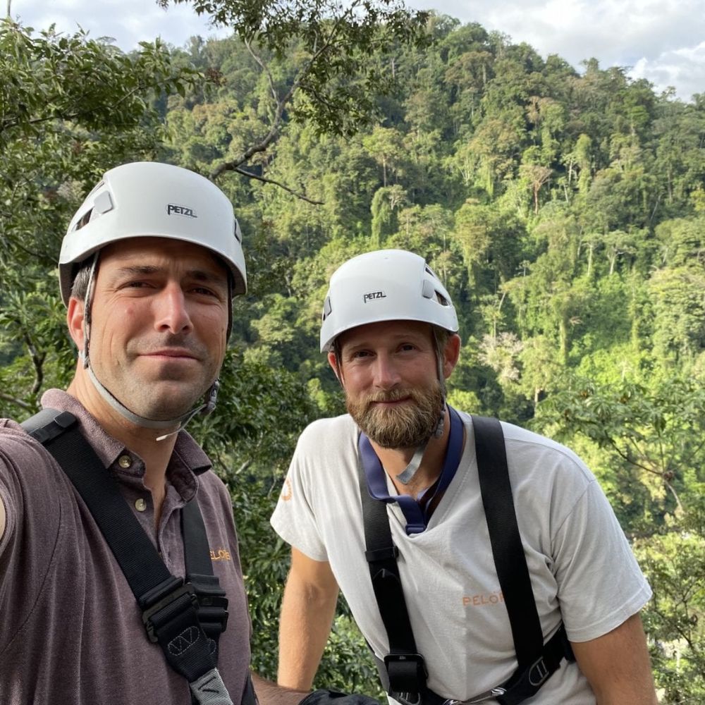 Two men wearing helmets and safety harnesses pose for a selfie in a lush, green forest. Tall trees and a distant hillside create a scenic backdrop. Both men are smiling at the camera.