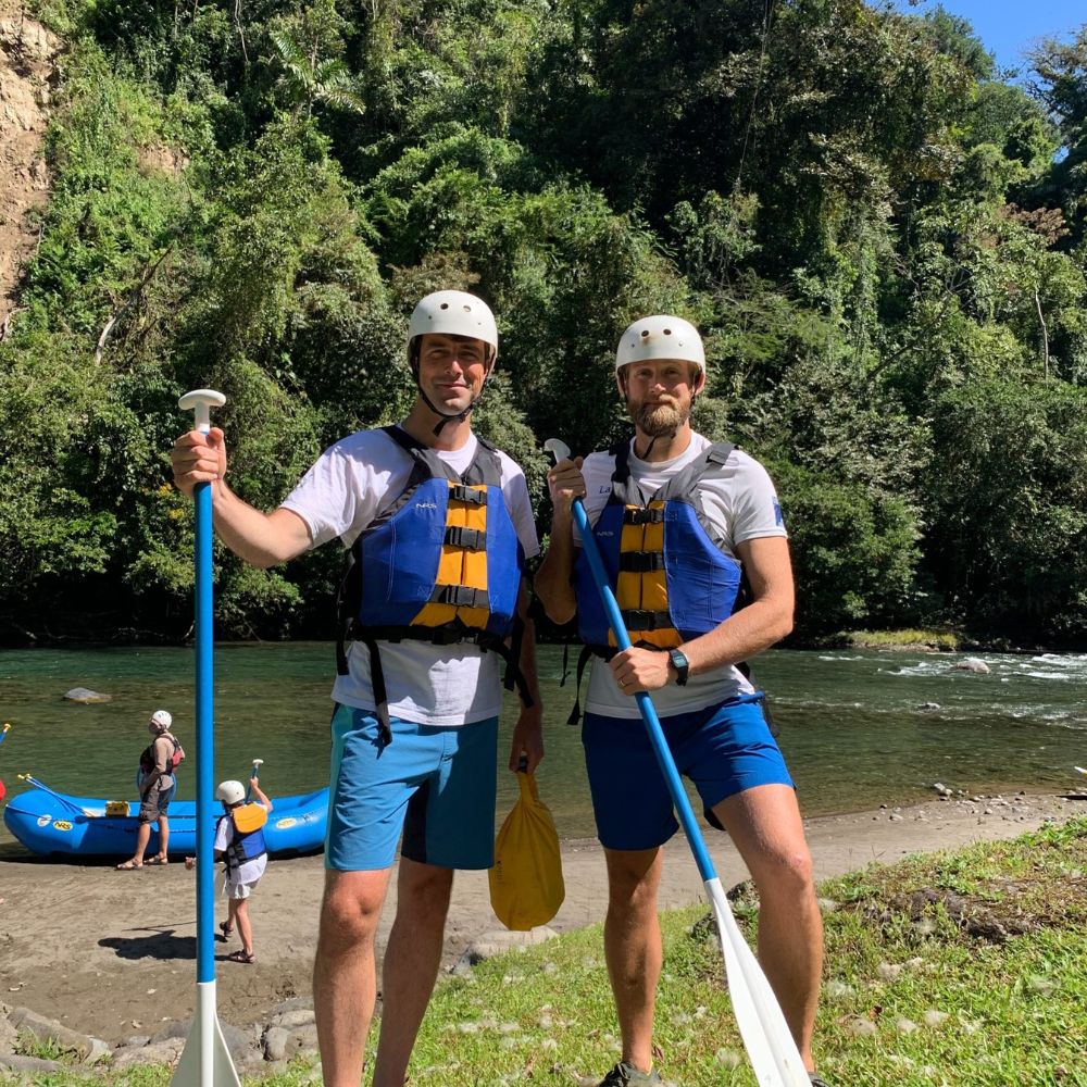 Two men stand on a grassy bank next to a river, each holding a paddle and wearing helmets and life jackets. Lush greenery surrounds them, with others in the background preparing for a rafting adventure.