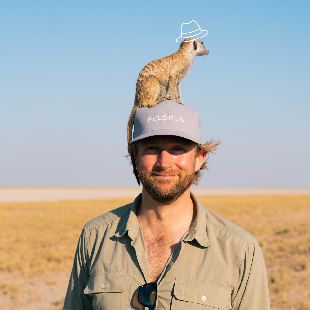 A man wearing a gray cap stands in a vast, dry landscape with a meerkat perched playfully on his head, which is topped with a cartoonish hat.
