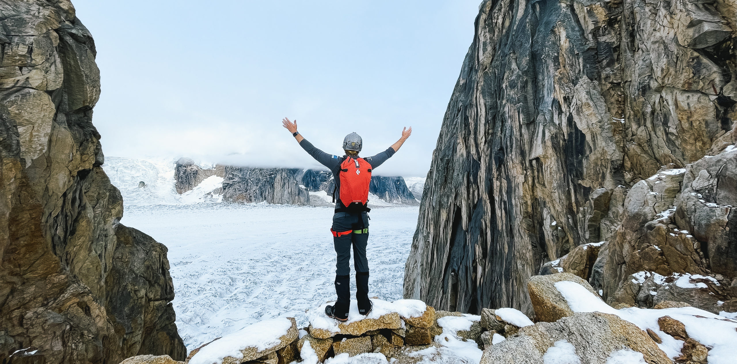 A climber stands on a rocky ledge with arms raised in triumph, surrounded by towering cliffs and a snowy landscape. Glaciers and mountains are visible in the background under a cloudy sky.