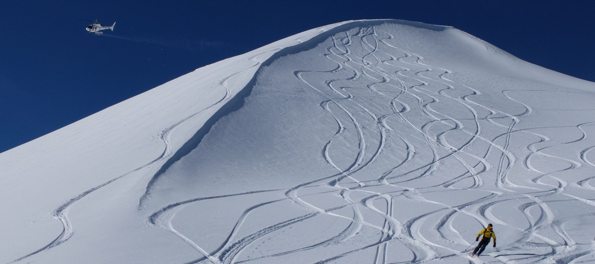 A skier descends a snow-covered mountain peak, leaving tracks in the fresh powder. A helicopter hovers above against a clear blue sky.