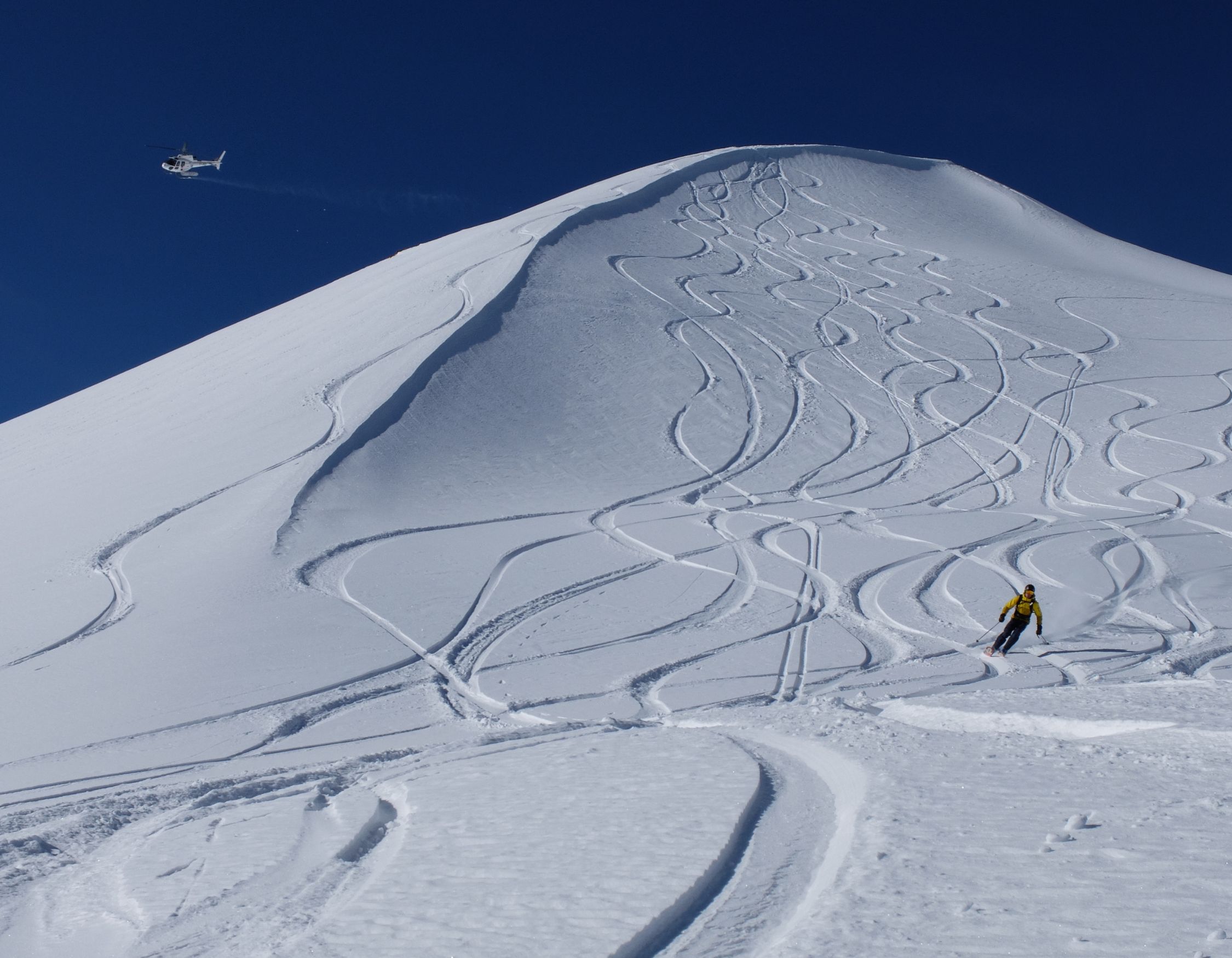 A skier descends a snowy mountain with distinct tracks in the fresh powder, while a helicopter hovers in the clear blue sky above.