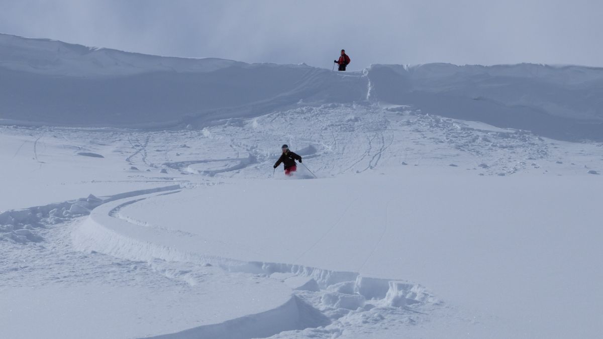 A skier descends a snowy slope, leaving deep tracks in fresh powder, while another skier stands on a ridge above, preparing to descend. Bright sunlight illuminates the scene against a cloudy sky.