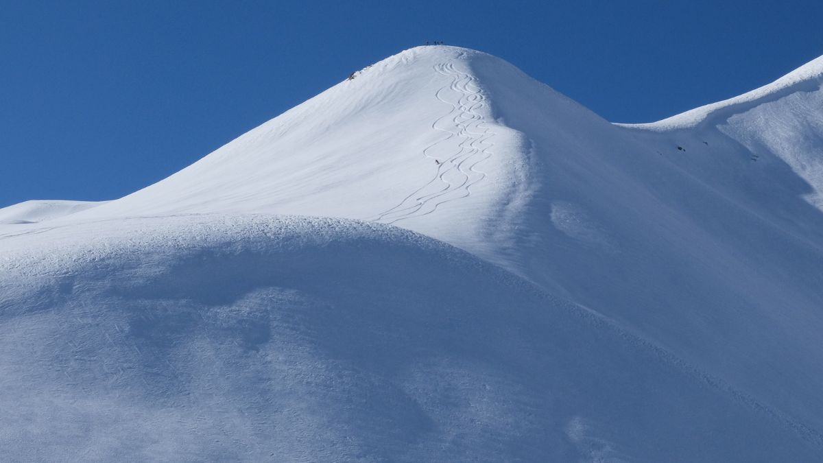 A pristine snow-covered mountain peak against a clear blue sky, with ski tracks leading up the slope and tiny figures near the summit.