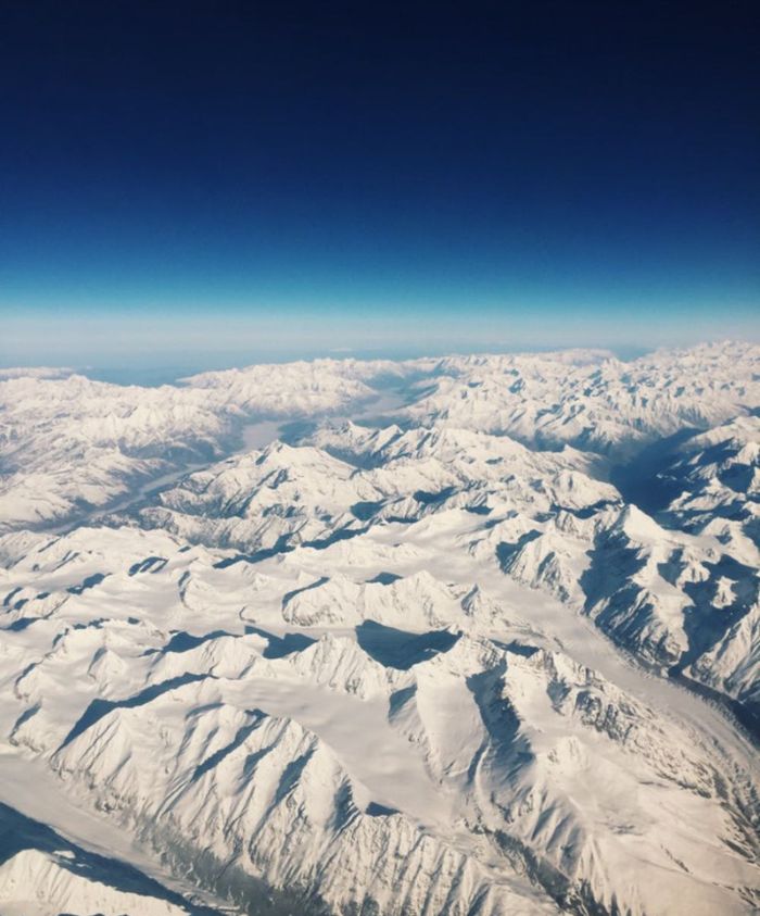 A breathtaking aerial view of rugged, snow-covered mountain peaks under a deep blue sky. The landscape features a mix of sharp ridges and smooth glaciers.