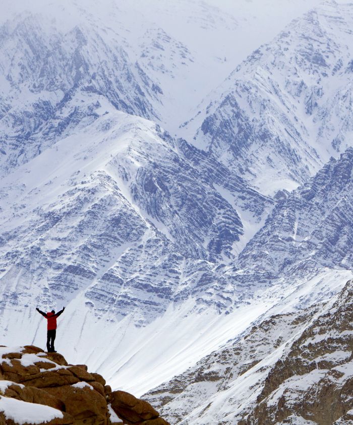 A person in a red jacket stands on a rocky outcrop with arms raised, surrounded by snow-covered mountains. The landscape features rugged peaks and a vast, snowy terrain under a cloudy sky.