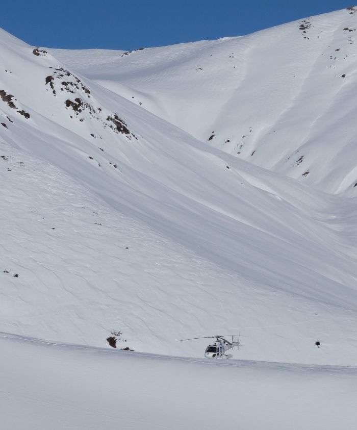A helicopter sits on a vast, snowy mountain landscape, with steep, snow-covered slopes and a clear blue sky in the background.