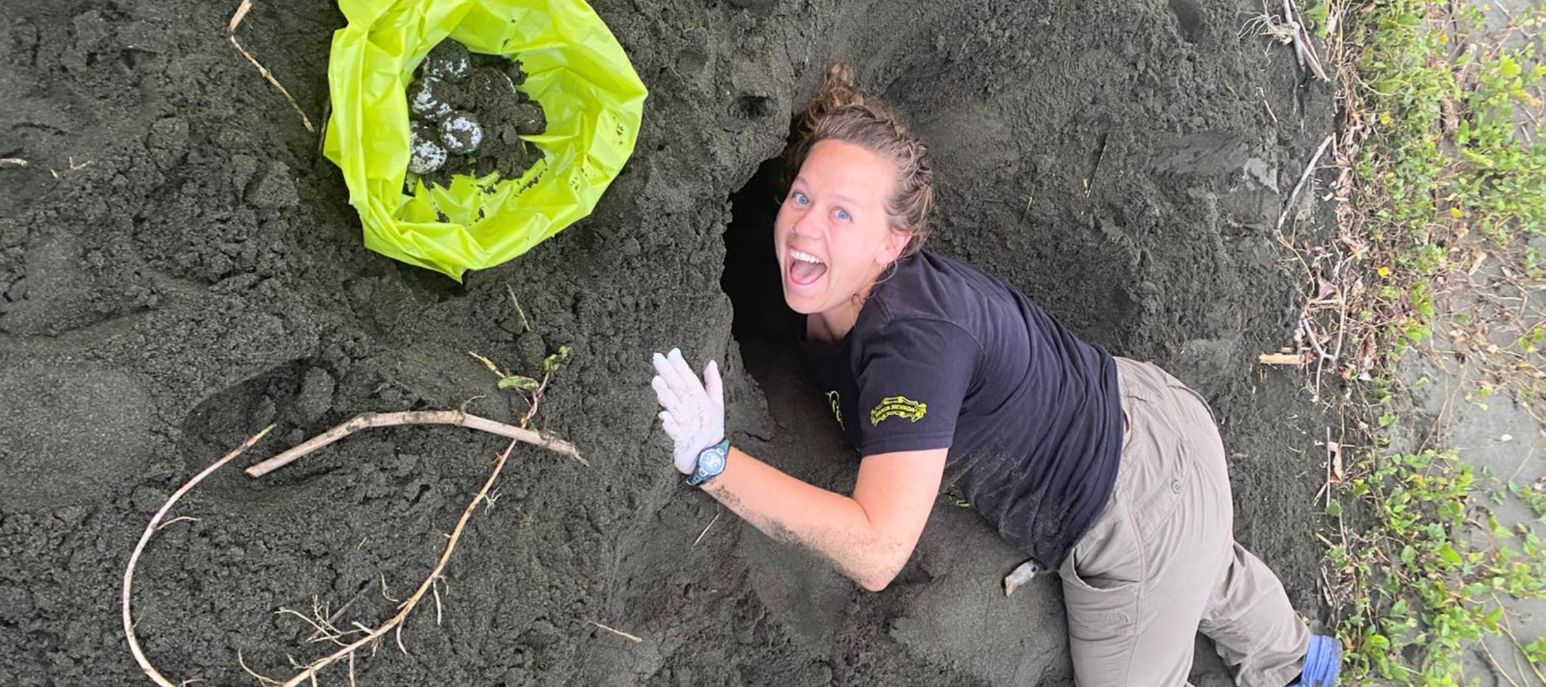 A woman is joyfully digging in dark sand on a beach, with a green bag beside her. She is wearing gloves and a black shirt, smiling at the camera.