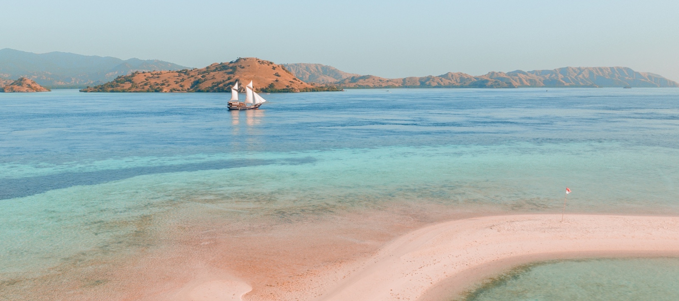 Aerial view of a traditional two-masted sailing ship anchored in turquoise water near a pink sandbar and tropical islands.