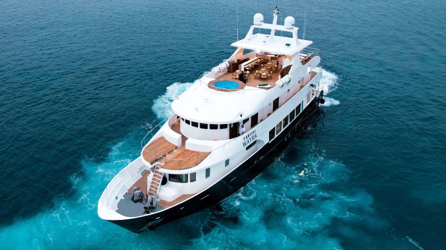 A large luxury yacht gliding over turquoise waters. The upper deck features a circular hot tub, and spacious outdoor seating areas. The yacht has multiple levels and a sleek design.