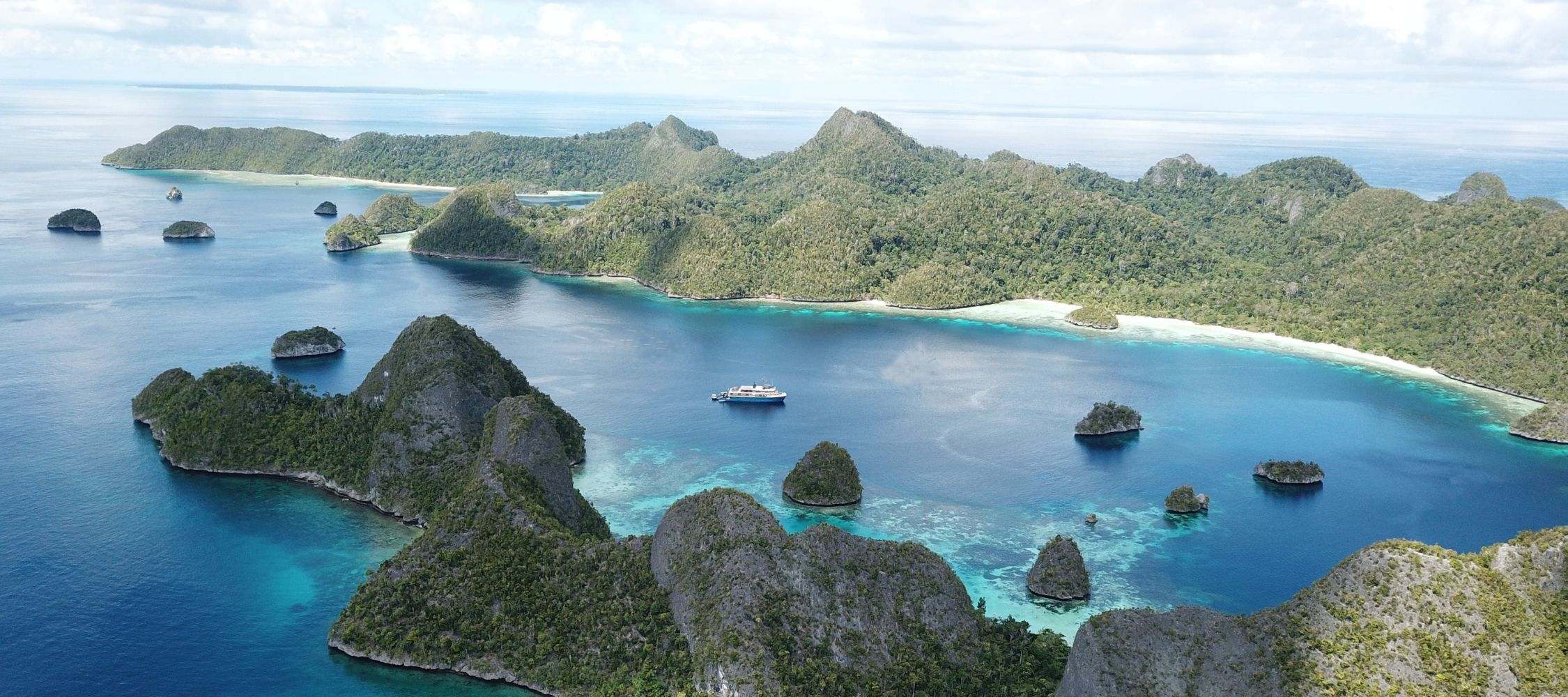 Aerial view of a small luxury ship anchored in a blue lagoon surrounded by lush, green limestone islands.