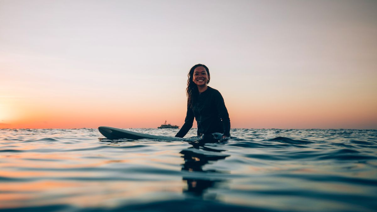 A Pelorus Yachting expert in a wetsuit sits on a surfboard in calm ocean water, smiling during a golden sunset.