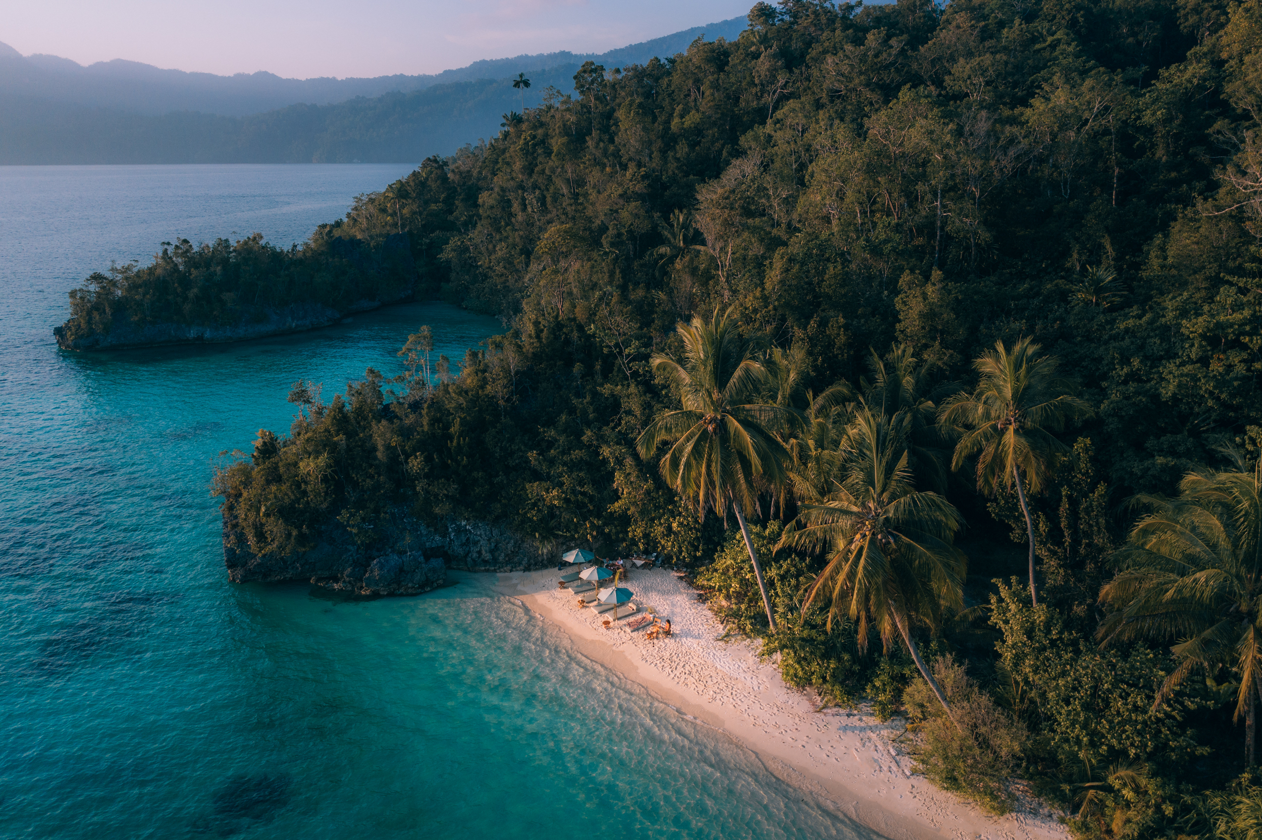 Aerial view of a secluded white sand beach with umbrellas, nestled between turquoise water and a dense tropical forest.