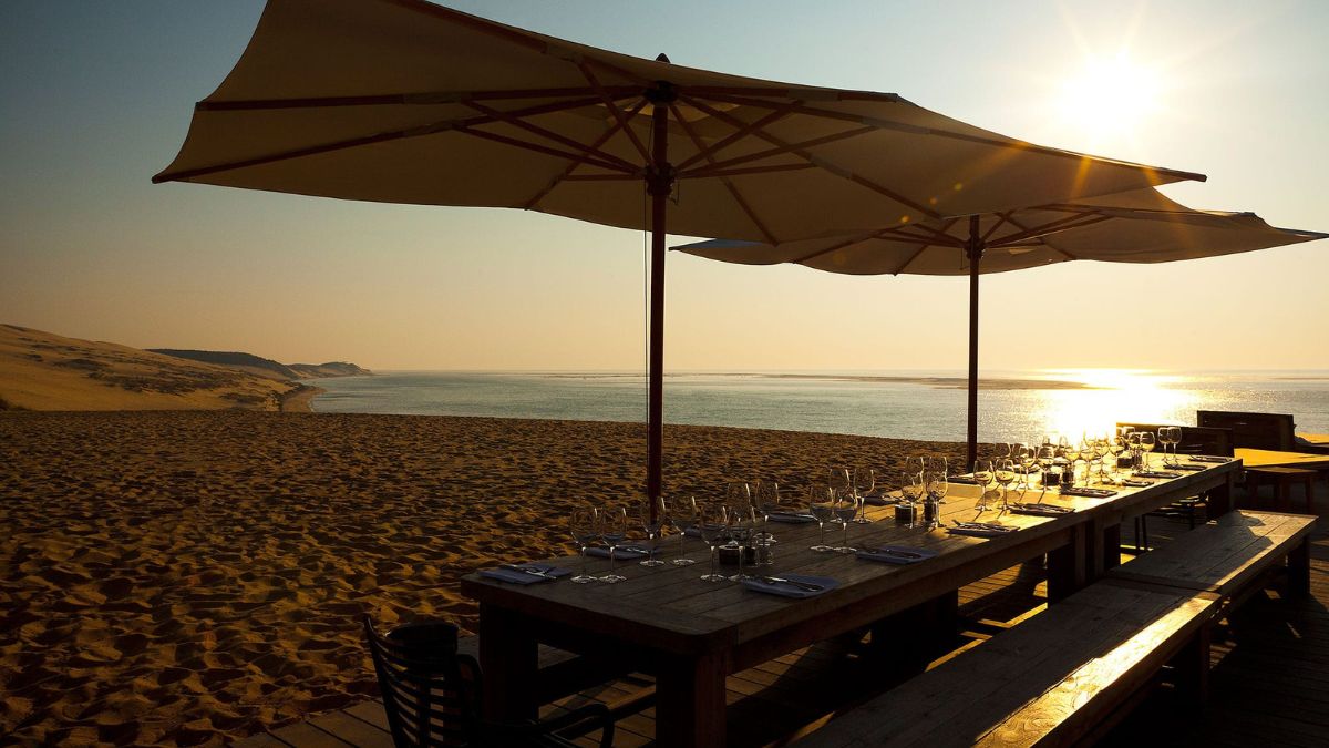 A beachfront dining setup featuring a long wooden table with glassware and plates, shaded by large umbrellas. The sun sets over the calm water, casting a warm glow on the sandy shore and distant dunes.