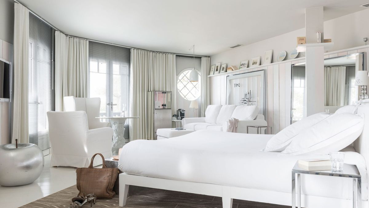 A bright, modern bedroom featuring white furniture, a spacious layout, and large windows with sheer curtains. A bed with fluffy white pillows is in the center, accompanied by a cozy seating area and a desk by the window. A stylish silver accent piece and a brown handbag add to the decor.