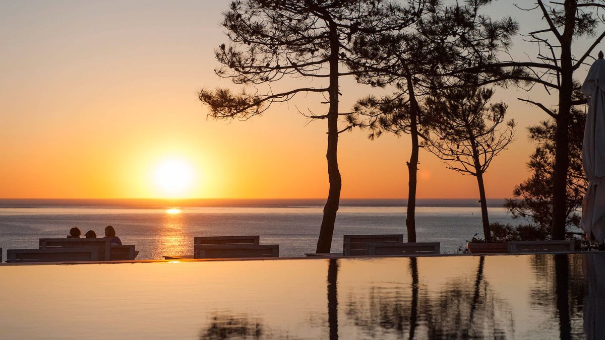 A serene sunset over the ocean, with silhouettes of trees and lounge chairs in the foreground. The sun casts a warm golden glow across the water, creating a tranquil atmosphere.