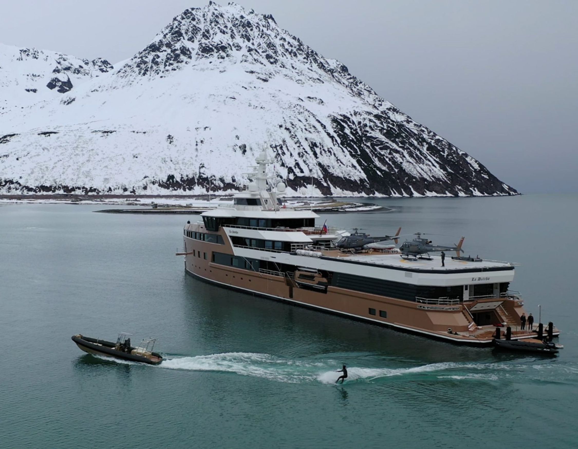 Luxury expedition yacht LA DATCHA in a snowy fjord with two helicopters on deck and a person wakeboarding behind a tender.