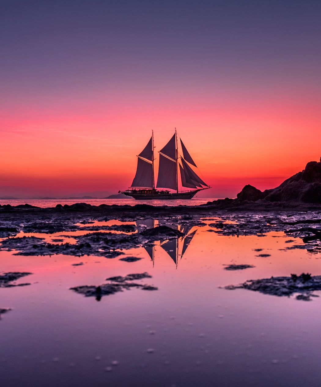Silhouette of a two-masted sailing ship on a calm sea, reflecting in a tide pool against a vibrant pink and orange sunset sky.