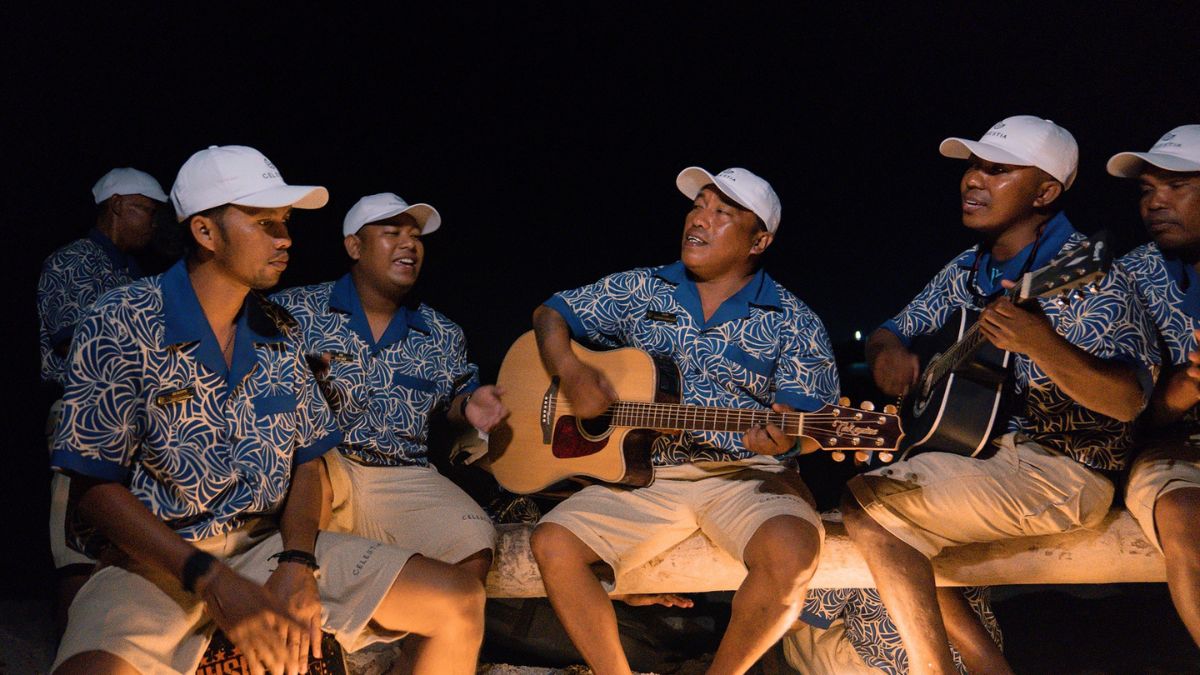 Group of men in matching blue patterned shirts and white caps playing acoustic guitars and singing at night.