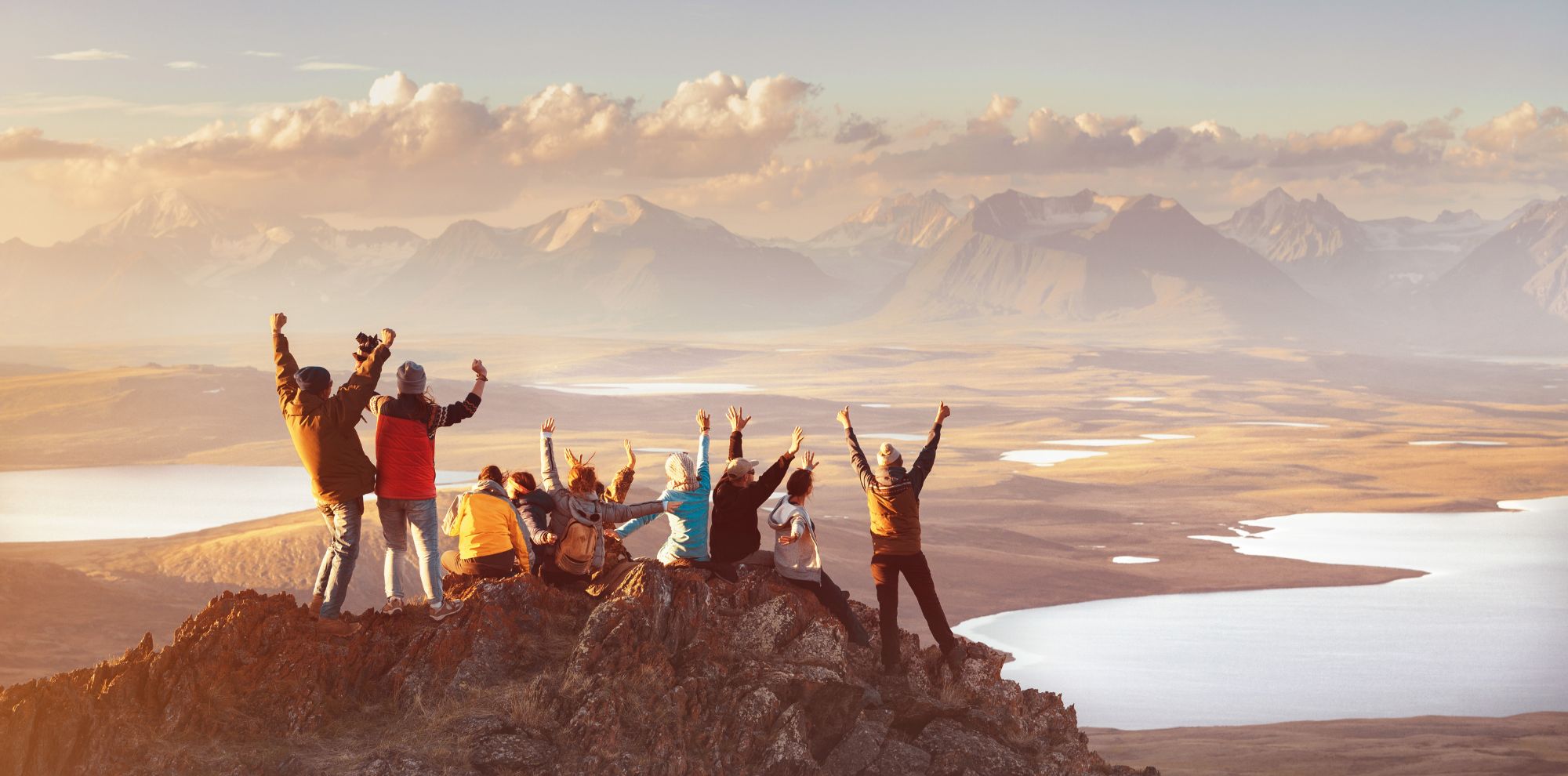 A group of hikers sitting on a rocky peak with their arms raised, overlooking a vast valley with lakes at sunset.