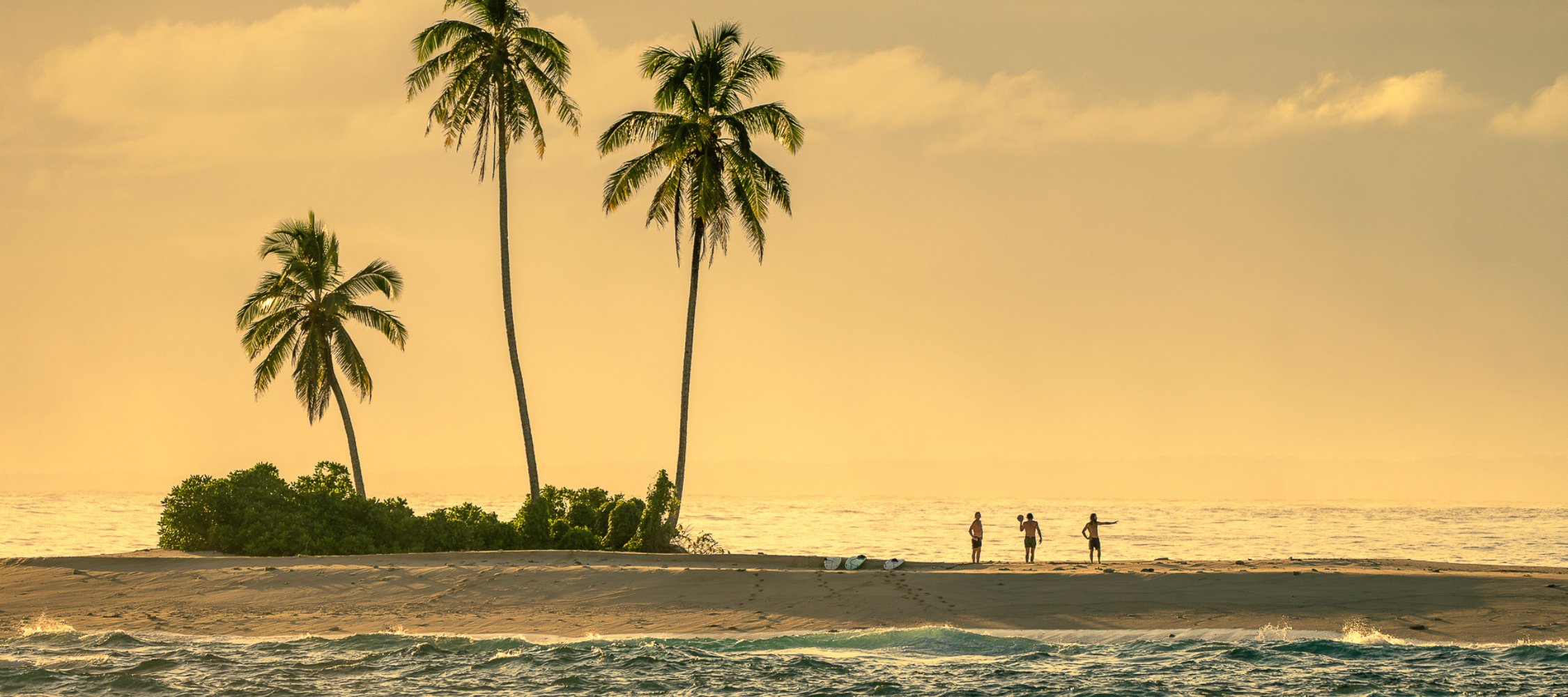 Three palm trees stand on a narrow sandy island at sunset, with three people standing on the shore.
