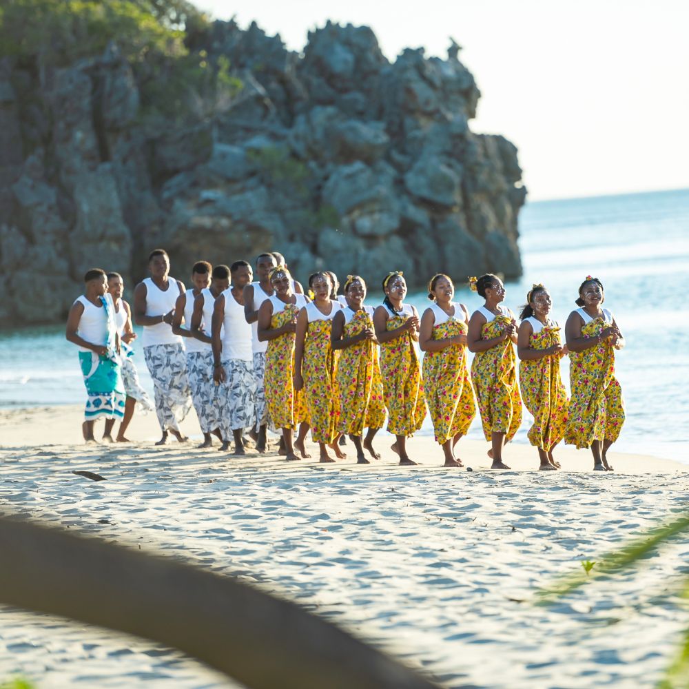 A group of people in traditional clothing, wearing floral patterns, walk along a sandy beach, with rocky cliffs and calm waters in the background. The scene evokes a festive atmosphere with smiles and unity among the participants.