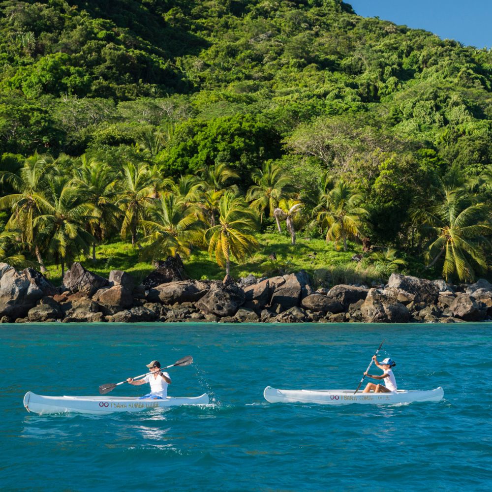Two people paddle in transparent kayaks on clear blue water, surrounded by lush greenery and palm trees along a rocky shoreline.