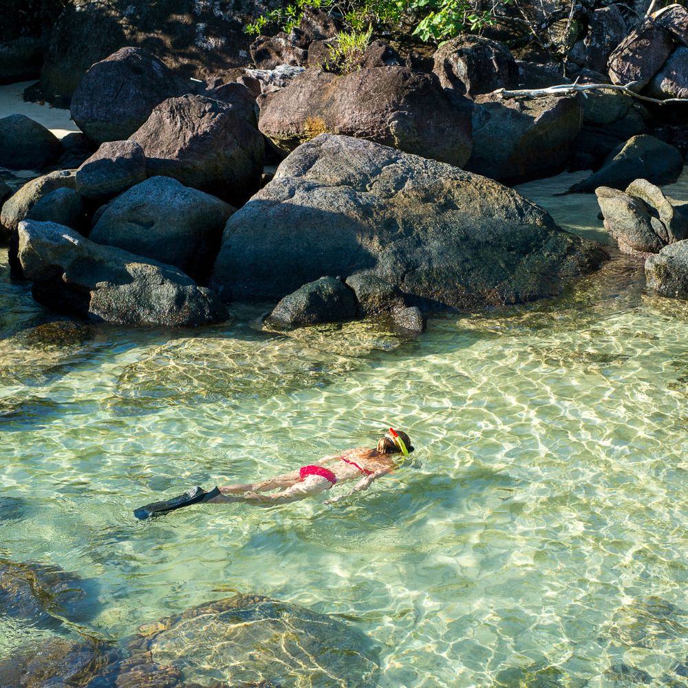 A person in a snorkel mask and fins swims in clear, shallow water surrounded by rocks. Sunny day with a tropical vibe.