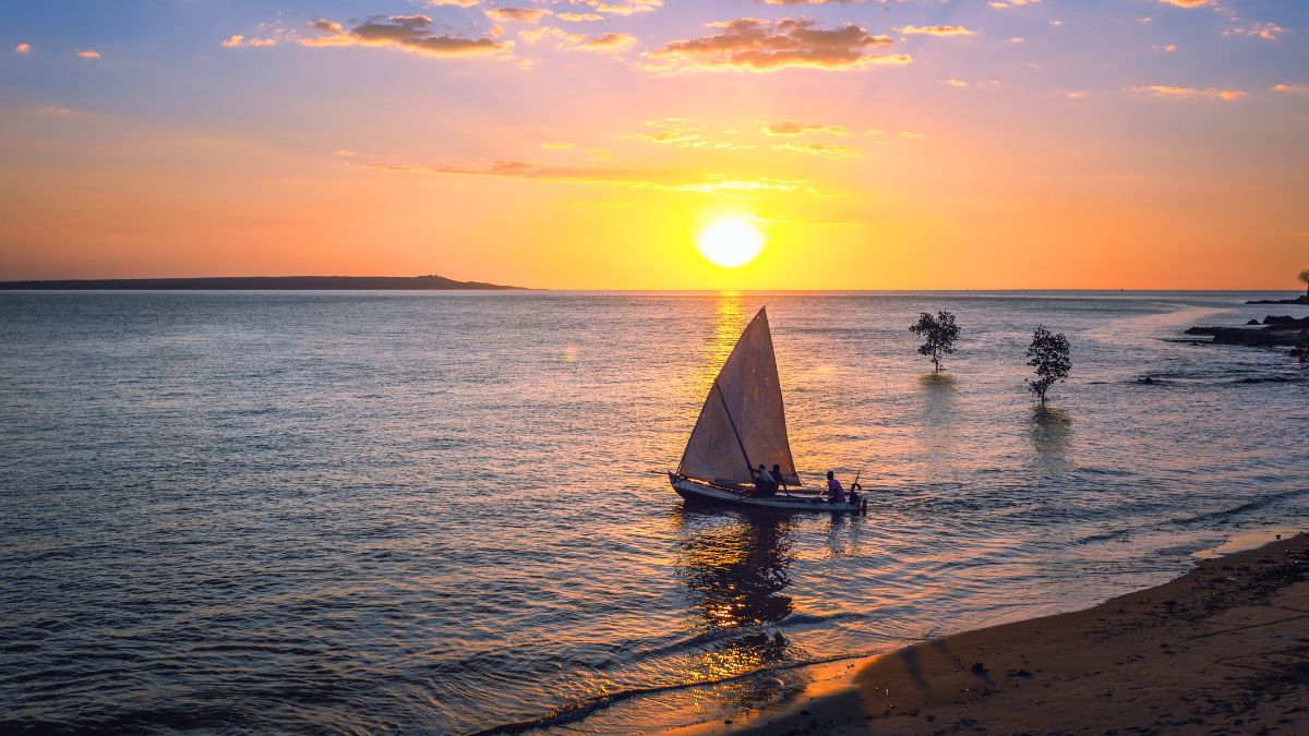 Silhouette of a sailboat with a triangular sail drifting on calm water during a bright orange sunset.