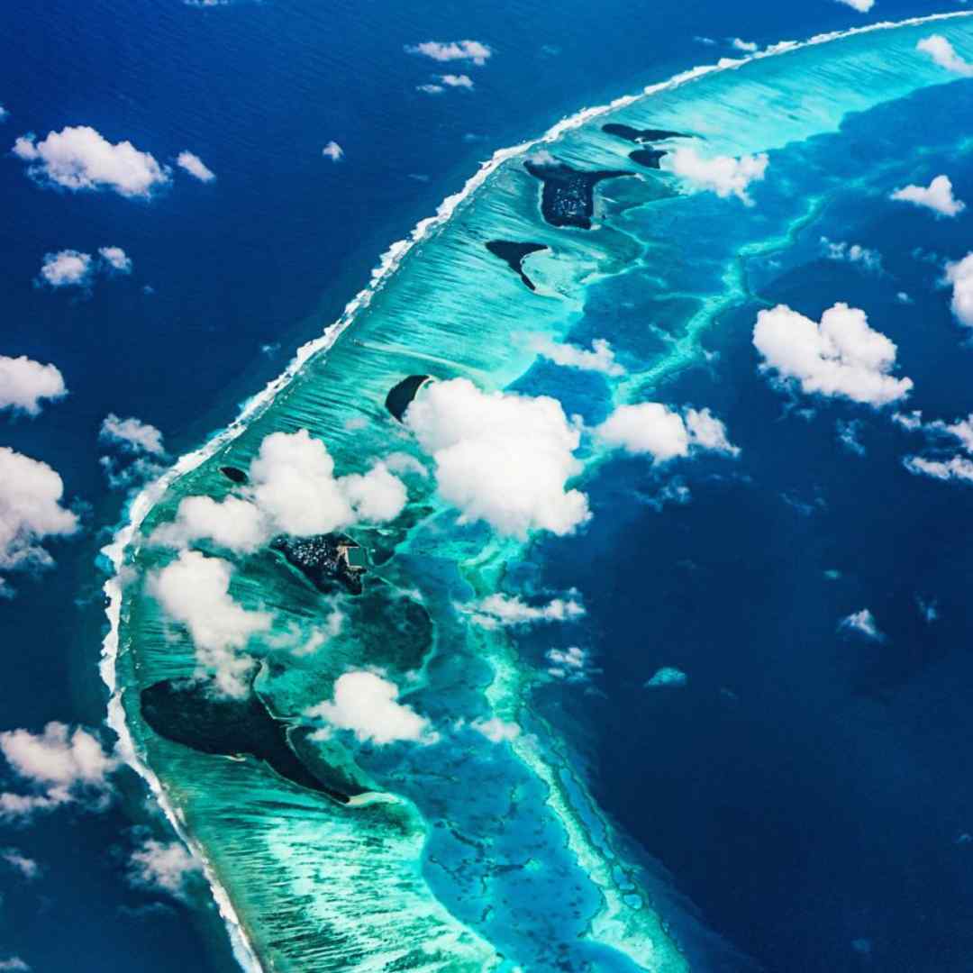 Aerial view of a stunning coral reef system with vibrant turquoise waters, surrounded by white sandy shores and scattered clouds above.