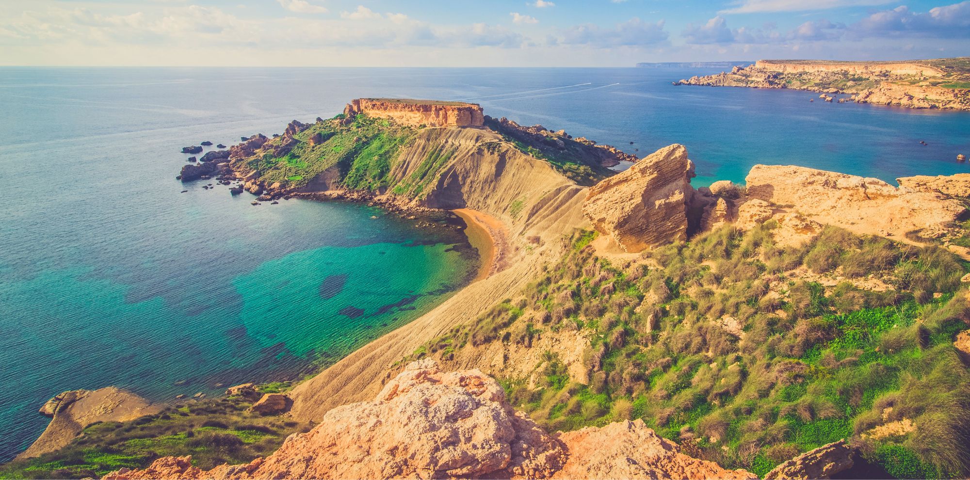 Wide shot of a rugged coastline with green hills, sandy coves, and turquoise water under a soft, hazy sky.