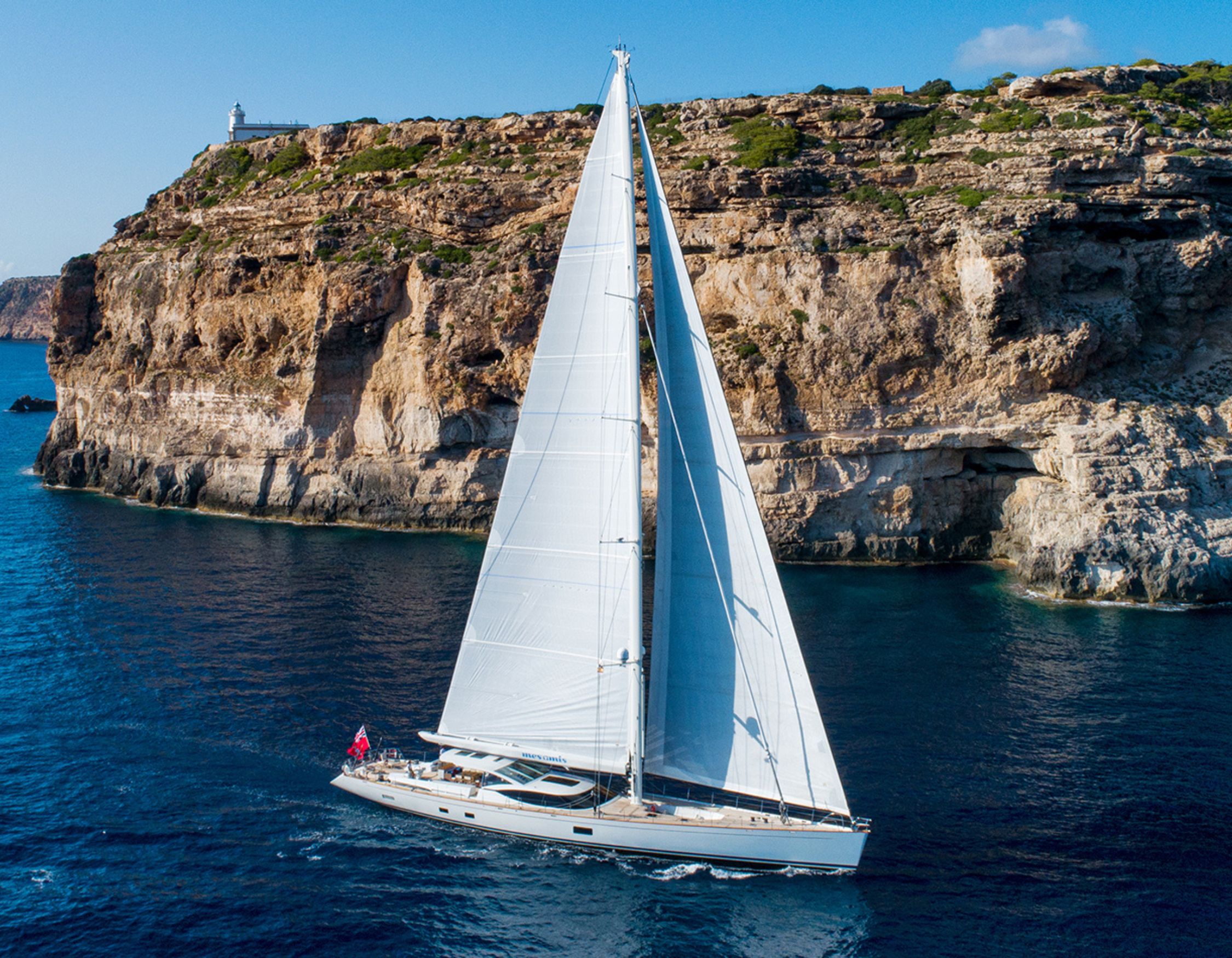 Large sailing yacht named MES AMIS with tall white sails gliding past a steep rocky cliff topped with a lighthouse.