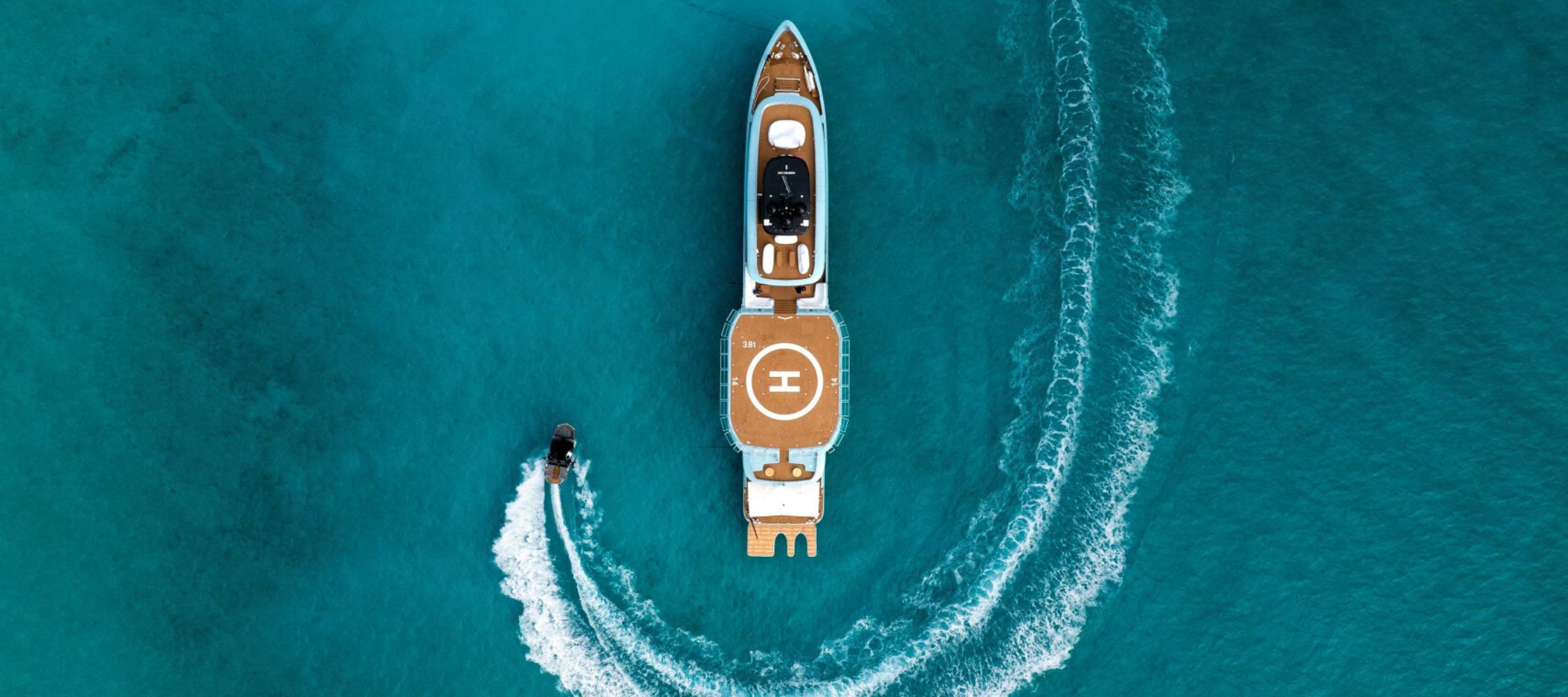 Blue luxury yacht with heli pad in clear blue water with a smaller boat driving around the boat.