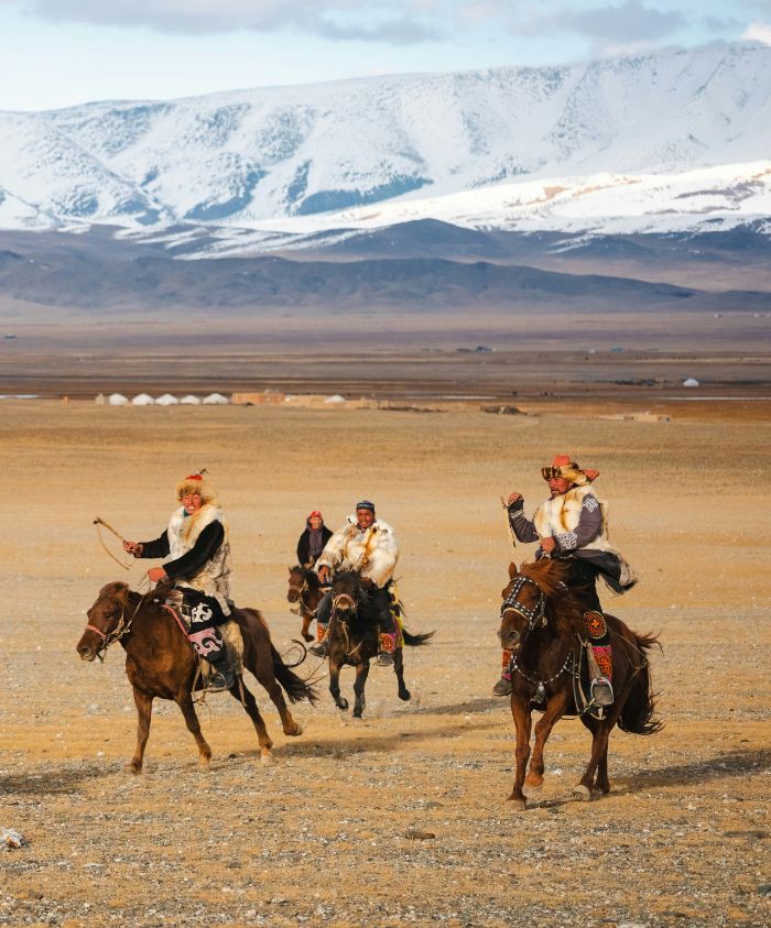 Mongolian eagle hunters riding horses across a vast plain toward snow-capped mountains.