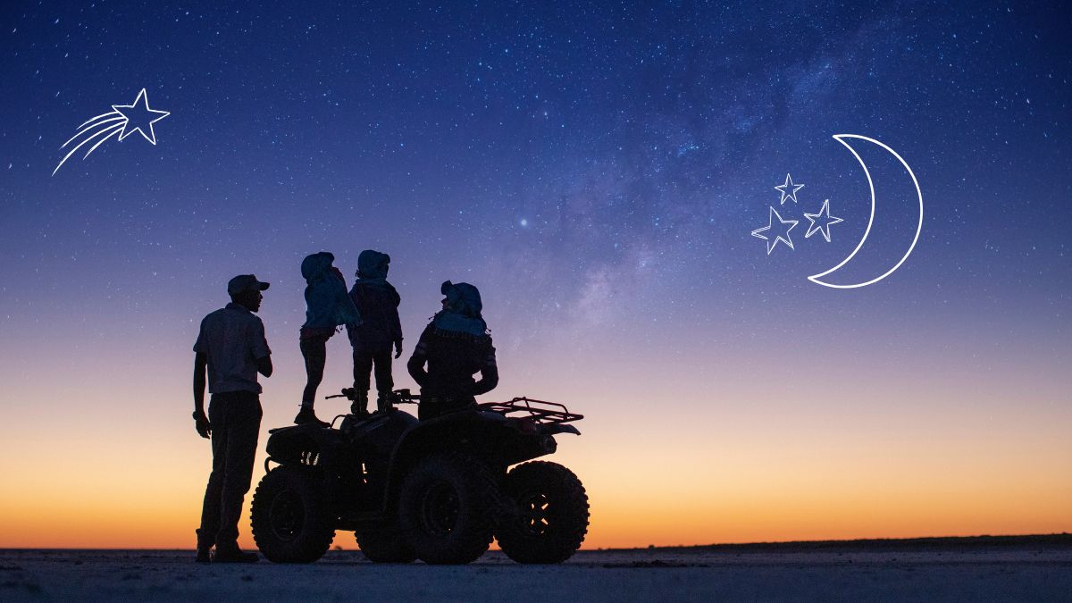 A family stands on an ATV, silhouetted against a twilight sky filled with stars and a visible Milky Way. Two children are perched atop the ATV, gazing at the horizon as the sun sets.
