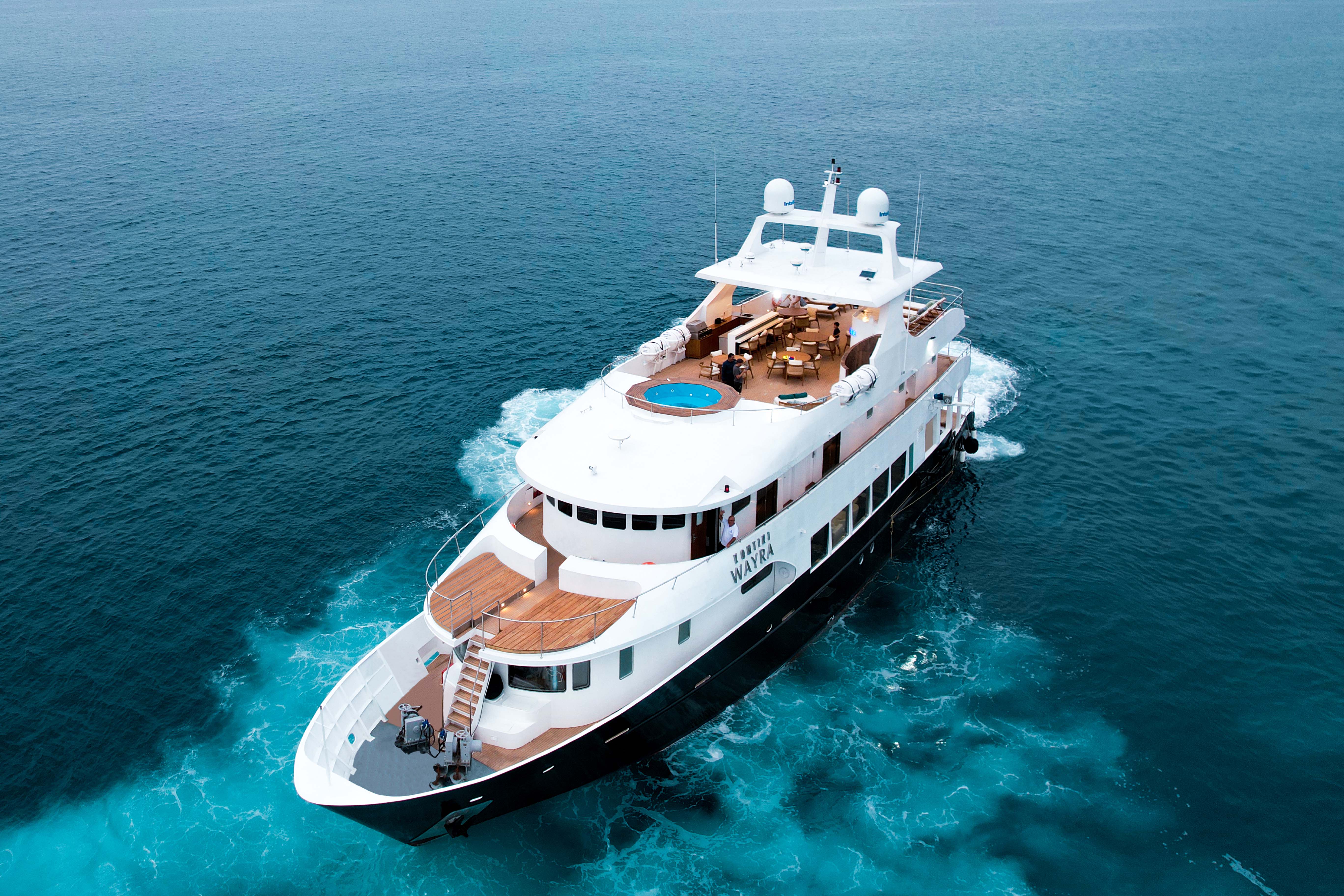 A luxurious yacht glides through turquoise waters, showcasing a spacious deck and a circular hot tub on top. The sleek white and dark hull contrasts beautifully with the ocean, highlighting its elegance.